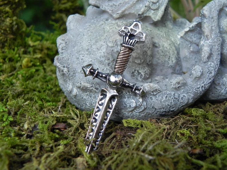 May include: A silver and copper sword pendant with intricate details. The sword has a pointed tip, a cross guard, and a twisted copper handle. The pendant is resting on a grey stone surface.