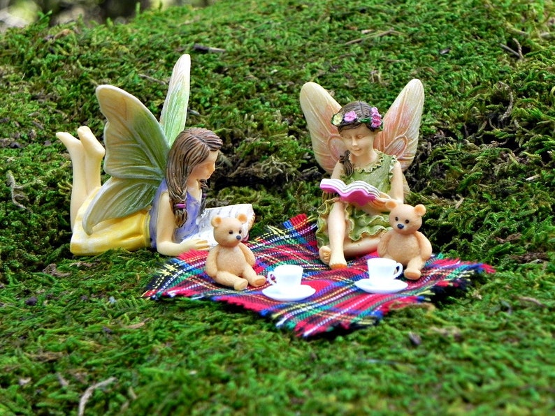 May include: Three fairies sitting on a plaid blanket with two teddy bears and two teacups. One fairy is reading a book, another is holding a book, and the third fairy is looking at the book being read.