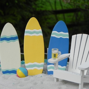 May include: A miniature beach scene with three colorful surfboards, a white Adirondack chair, and a beach ball. The surfboards are painted with blue and white waves. The chair is made of white wood and has a glass of beer on the armrest. The beach ball is yellow, blue, and white.