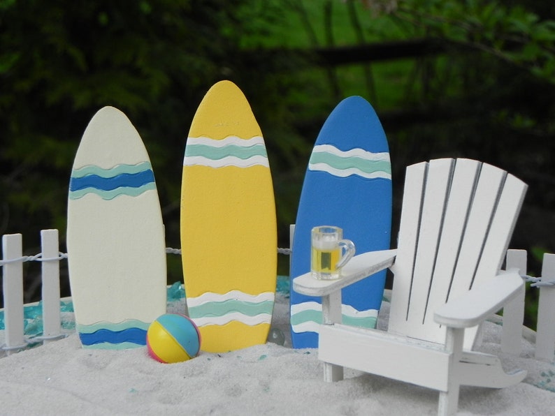 May include: A miniature beach scene with three colorful surfboards, a white Adirondack chair, and a beach ball. The surfboards are painted with blue and white waves. The chair is made of white wood and has a glass of beer on the armrest. The beach ball is yellow, blue, and white.