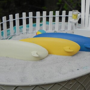 May include: Three miniature surfboards in a white sand beach scene. The surfboards are yellow, white, and blue. The scene is set in a white plastic container with a white picket fence and a miniature beach chair.