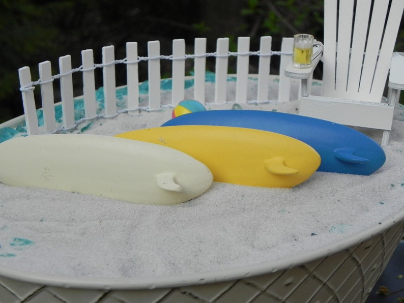May include: Three miniature surfboards in a white sand beach scene. The surfboards are yellow, white, and blue. The scene is set in a white plastic container with a white picket fence and a miniature beach chair.