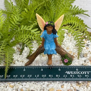 May include: A miniature fairy figurine with a blue dress, seated on a brown log. The fairy has tan wings and a floral crown. The scene includes green foliage and white pebbles. A ruler shows measurements in inches.