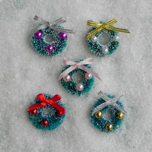 May include: Five miniature Christmas wreaths with different colored bows and beads. The wreaths are green and made of a fuzzy material. The bows are pink, red, silver, and gold.