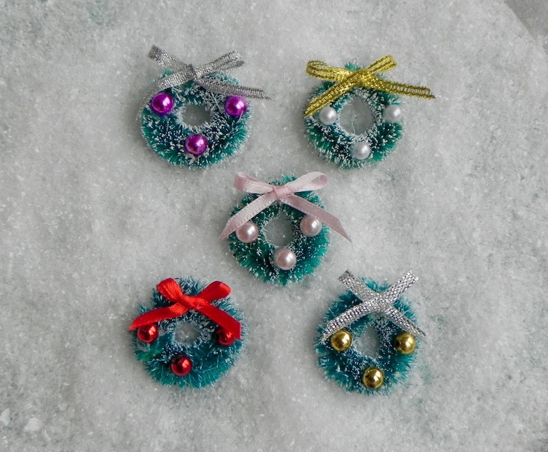 May include: Five miniature Christmas wreaths with different colored bows and beads. The wreaths are green and made of a fuzzy material. The bows are pink, red, silver, and gold.