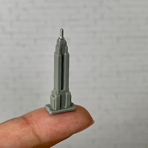 May include: A miniature gray Empire State Building model with a silver antenna. The building is held on a fingertip, showcasing its small size. The model is a detailed replica of the iconic skyscraper.