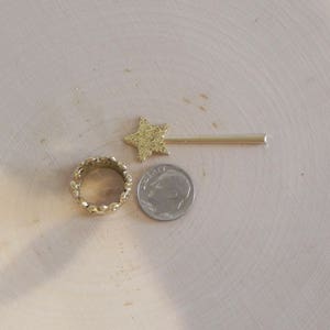 May include: A gold star-shaped hair clip, a gold ring with a textured design, and a silver coin with the inscription "Liberty" on a light wood surface.