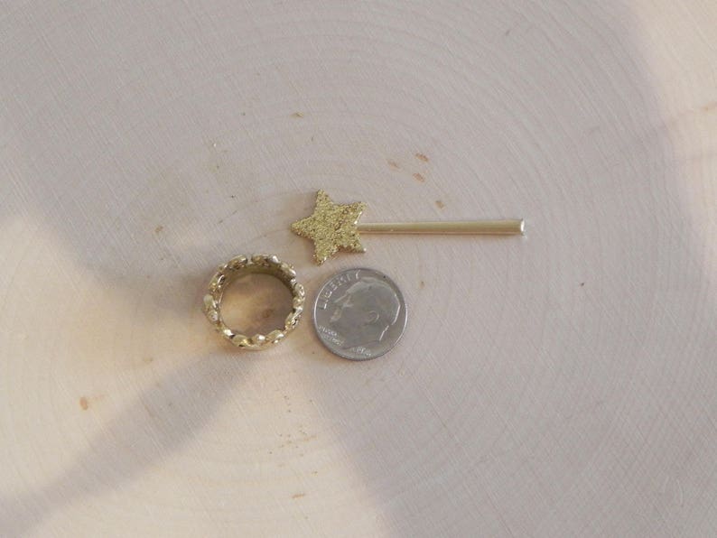 May include: A gold star-shaped hair clip, a gold ring with a textured design, and a silver coin with the inscription "Liberty" on a light wood surface.