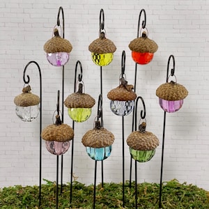 May include: Ten miniature acorn-shaped glass lanterns on black metal stakes. The lanterns are filled with colorful glass beads and are arranged in a row on a bed of green moss.