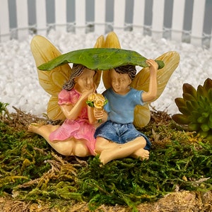 May include: A miniature fairy garden figurine featuring a boy and girl under a leaf. The girl wears a pink dress and holds flowers, while the boy wears a blue shirt and denim shorts. The figurines have yellow wings and are set on a bed of moss.