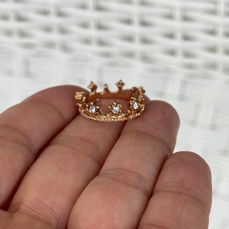 May include: A gold crown ring with clear rhinestones. The ring is on a hand.