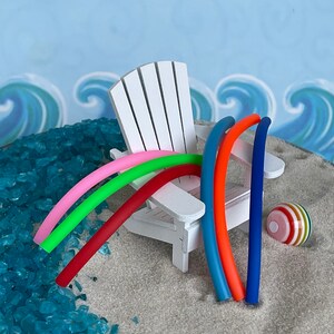 May include: A white miniature Adirondack chair with five colourful pool noodles leaning against it. A striped beach ball sits on the sand next to the chair.