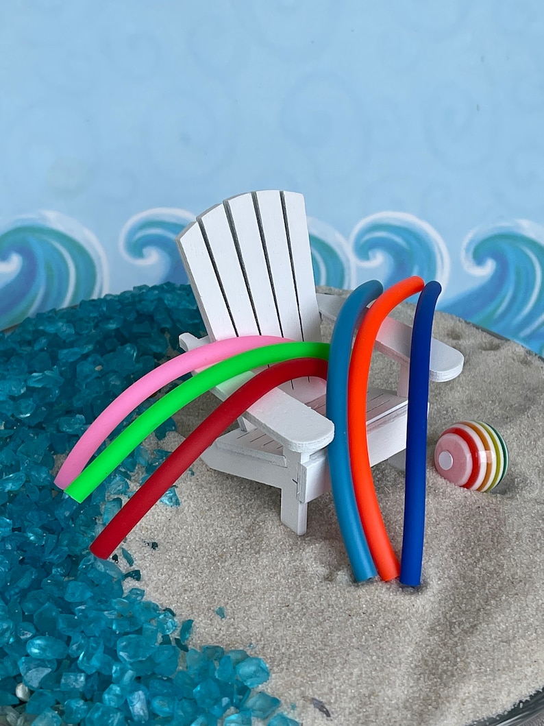 May include: A white miniature Adirondack chair with five colourful pool noodles leaning against it. A striped beach ball sits on the sand next to the chair.