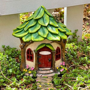 May include: A whimsical fairy house with a leaf-covered roof and a red door that reads "Fairies Welcome." The house has small windows, a stone path, and is surrounded by greenery and flowers. The house is made of a light brown material.