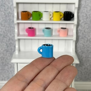 May include: A miniature blue ceramic coffee mug with a black rim and handle. The mug is sitting on a hand in front of a white hutch with shelves filled with colorful mugs.