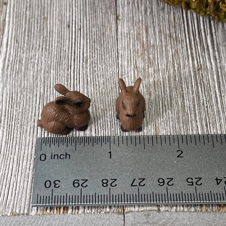 May include: Two small brown rabbit figurines. One is posed lying down, the other is upright. A ruler is placed next to the figurines, showing measurements in inches. The rabbits are made of a solid material and have detailed features.