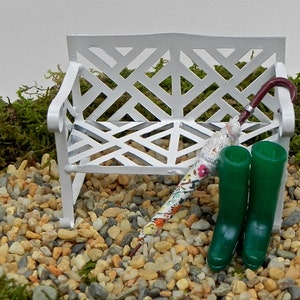May include: A miniature garden scene featuring a white bench with a lattice design, a colorful floral umbrella, and a pair of green rain boots. The scene is set on a bed of small pebbles and moss.