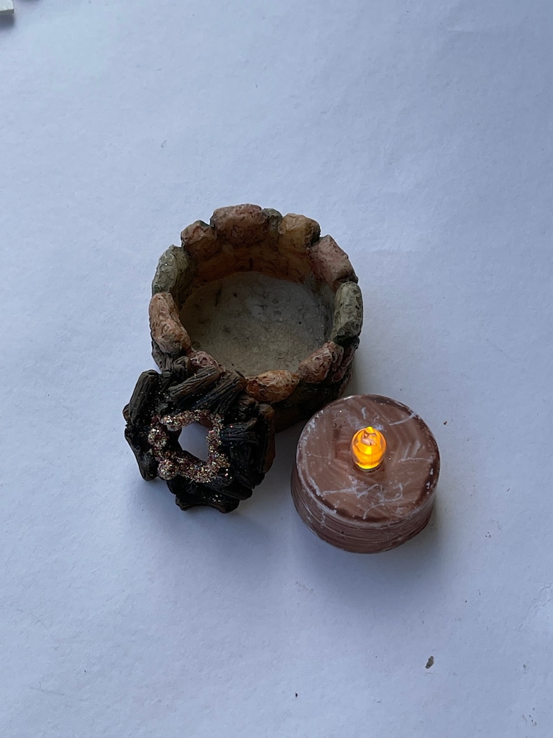 K&ouml;nnte beinhalten: Ein Miniatur-Steinkamin mit einem braunen und schwarzen Holzring und einer kleinen, runden, braunen Kerze mit einem warmen, orangenen Schein.