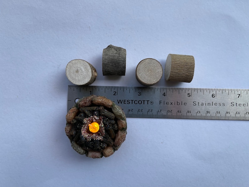 K&ouml;nnte beinhalten: Ein Miniatur-Lagerfeuer mit einem gl&uuml;henden orangefarbenen Licht in der Mitte, umgeben von kleinen braunen und grauen Holzscheiten. Es gibt vier weitere Holzscheite, zwei braune und zwei graue, die um ein Lineal angeordnet sind.