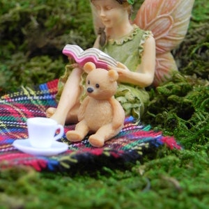 May include: A miniature fairy figurine reading a book, with a teddy bear and a teacup on a plaid blanket. The fairy has wings and a flower crown. The scene is set on a bed of green moss.