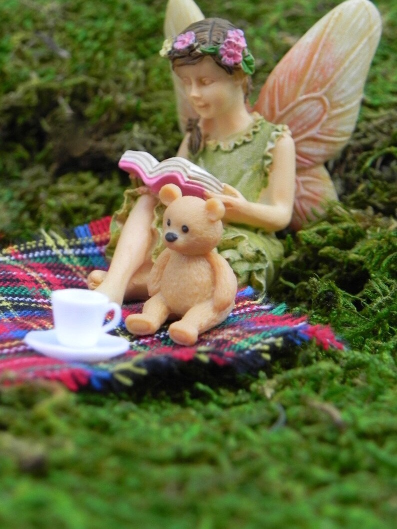May include: A miniature fairy figurine reading a book, with a teddy bear and a teacup on a plaid blanket. The fairy has wings and a flower crown. The scene is set on a bed of green moss.
