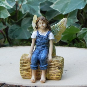 May include: A miniature fairy figurine with brown hair, wearing blue denim overalls and a white shirt, sits on a yellow hay bale. The fairy has yellow and green wings and is barefoot. The figurine is a decorative garden ornament.