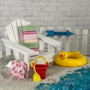 May include: A miniature white adirondack chair with a pink and green striped towel, a pink and white flip-flop, a red bucket and shovel, and a yellow rubber duck ring float. The chair is in front of a white picket fence with a blue sign that says "BEACH".