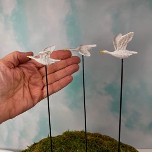 May include: Three white goose garden stakes with yellow beaks and black stems are displayed against a mossy green mound and a blue and white sky background. The geese are in various stages of flight.