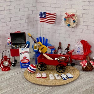 May include: Miniature scene with a red wagon, grill, and Adirondack chair, evoking a backyard barbecue. Includes a guitar, American flag, and a bag of charcoal. A festive wreath and a red and white striped ball add to the patriotic theme.