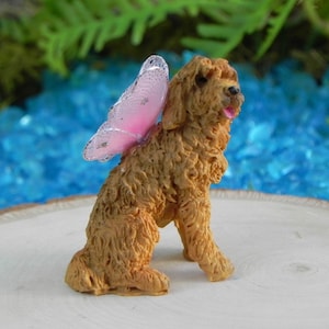 May include: A small, brown dog figurine with pink butterfly wings. The dog is sitting with its tongue out, and the wings are attached to its back. The figurine is on a wooden surface with a blue and green background.