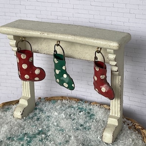May include: A miniature Christmas mantelpiece with three decorative stockings. The stockings are red and green with white polka dots. The mantelpiece is off-white and sits above a bed of faux snow.