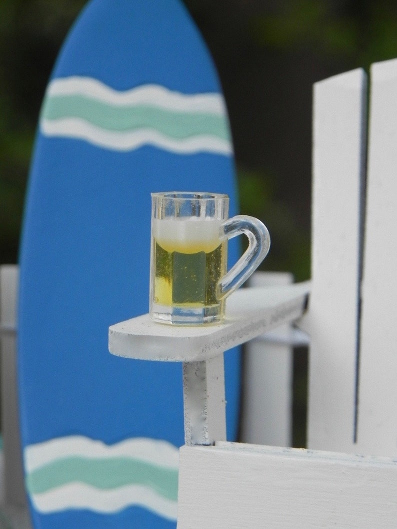 May include: A white wooden chair with a glass of beer on the armrest. The chair is in front of a blue and white surfboard.