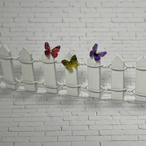 May include: A white picket fence with three butterflies attached to it. The butterflies are purple, yellow, and pink.