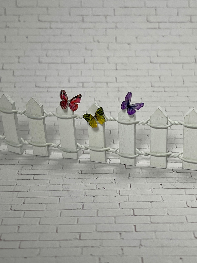 May include: A white picket fence with three butterflies attached to it. The butterflies are purple, yellow, and pink.