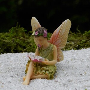 May include: A miniature fairy figurine, seated and reading a book. The fairy wears a green dress and a floral crown. Her wings are a soft peach color. The figurine is set on a bed of small white stones, with a blurred green background.