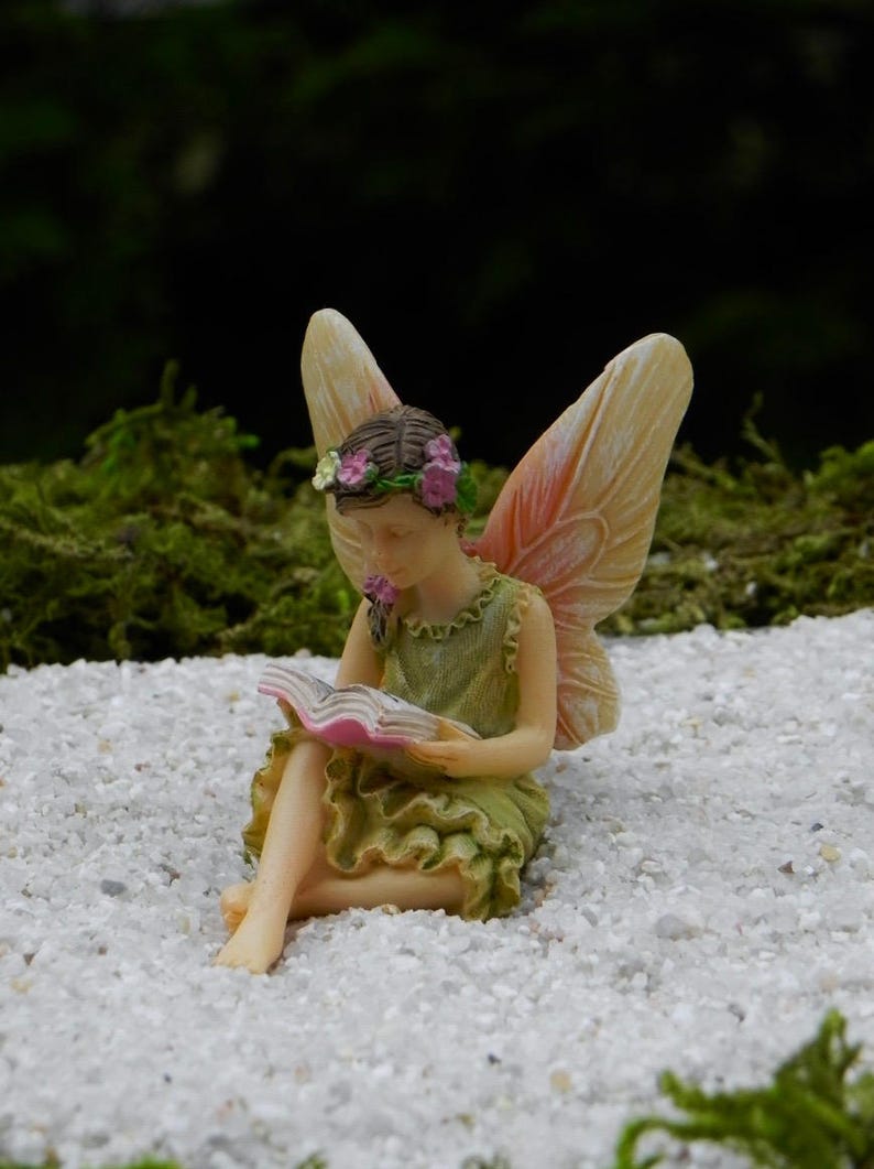 May include: A miniature fairy figurine, seated and reading a book. The fairy wears a green dress and a floral crown. Her wings are a soft peach color. The figurine is set on a bed of small white stones, with a blurred green background.