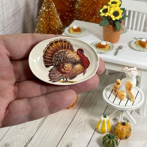 May include: A miniature oval plate featuring a detailed illustration of a turkey in shades of brown, red, and cream. The plate is held in a hand, with a miniature Thanksgiving scene in the background, including pumpkins, a cat, and a table setting.