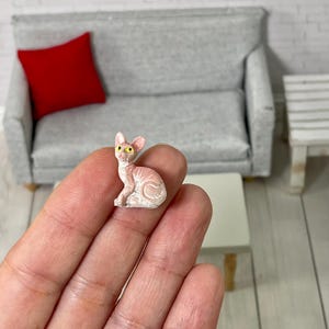 May include: A small, pink, hairless cat figurine with yellow eyes. The figurine is sitting on a hand.