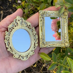 May include: Two ornate gold-toned framed mirrors, one round and one square, with a reflective surface. The mirrors are held in a hand against a green leafy background.