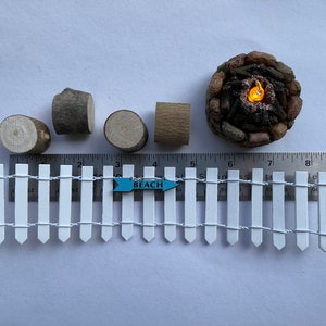 K&ouml;nnte beinhalten: Ein Miniatur-Lattenzaun in Wei&szlig; mit einem blauen Schild, auf dem "BEACH" steht, und ein Miniatur-Lagerfeuer mit einer gl&uuml;henden orangefarbenen Flamme. In der N&auml;he befinden sich auch vier Miniatur-Holzscheite.