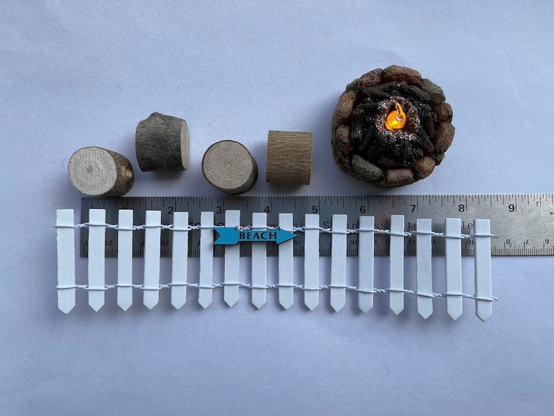 K&ouml;nnte beinhalten: Ein Miniatur-Lattenzaun in Wei&szlig; mit einem blauen Schild, auf dem "BEACH" steht, und ein Miniatur-Lagerfeuer mit einer gl&uuml;henden orangefarbenen Flamme. In der N&auml;he befinden sich auch vier Miniatur-Holzscheite.