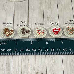 May include: Six miniature Christmas-themed plates with various decorations, including reindeer, gingerbread men, snowmen, and stockings. The plates are arranged on a wooden surface with a ruler for scale.