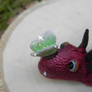 May include: A small red dragon figurine with a green and white butterfly on its head. The dragon has a detailed textured surface and a single large eye.