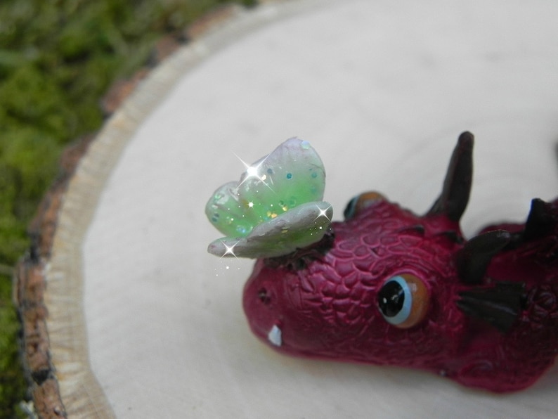 May include: A small red dragon figurine with a green and white butterfly on its head. The dragon has a detailed textured surface and a single large eye.