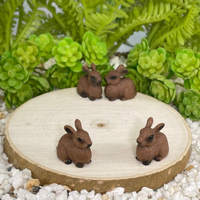 May include: Four small, brown rabbit figurines are arranged on a light wood slice, surrounded by white pebbles and green foliage. The rabbits are in various poses, creating a miniature woodland scene.