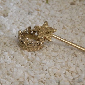 May include: A gold crown and a gold wand with a glitter star on top. The wand is lying on a white surface.