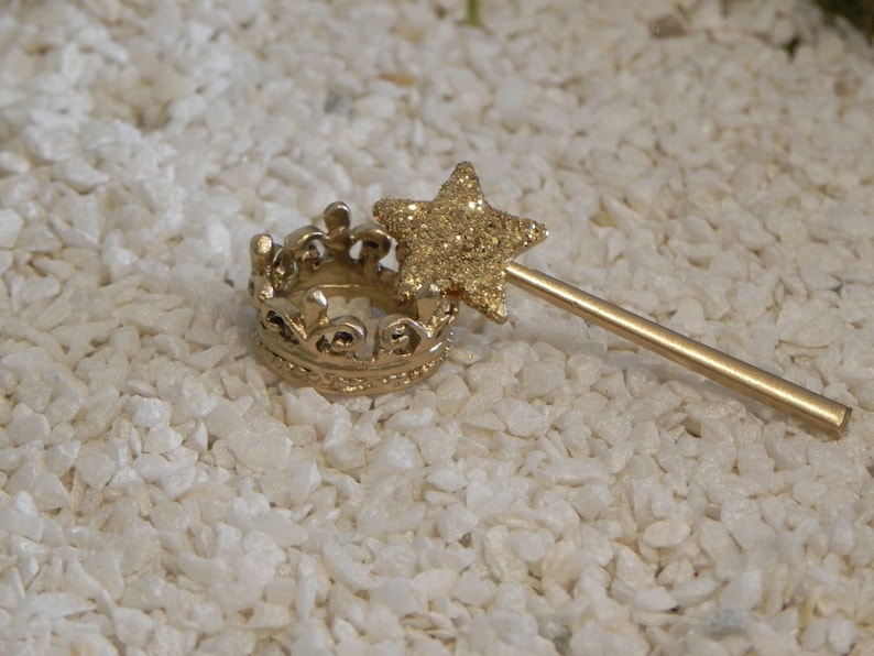 May include: A gold crown and a gold wand with a glitter star on top. The wand is lying on a white surface.