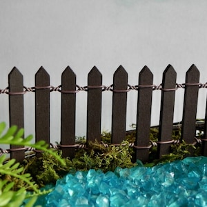May include: A miniature brown picket fence with a wire top, surrounded by green moss and blue glass stones.