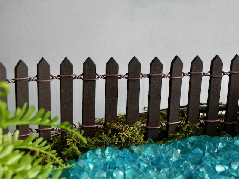 May include: A miniature brown picket fence with a wire top, surrounded by green moss and blue glass stones.