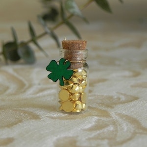 May include: A small glass bottle filled with gold-colored, faceted beads. The bottle is sealed with a cork and adorned with a green shamrock charm. The bottle is approximately 2 inches tall.